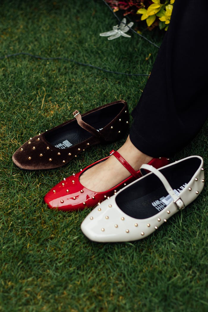 Three studded flats in red, brown, and white placed on grass, showcasing fashion footwear.
